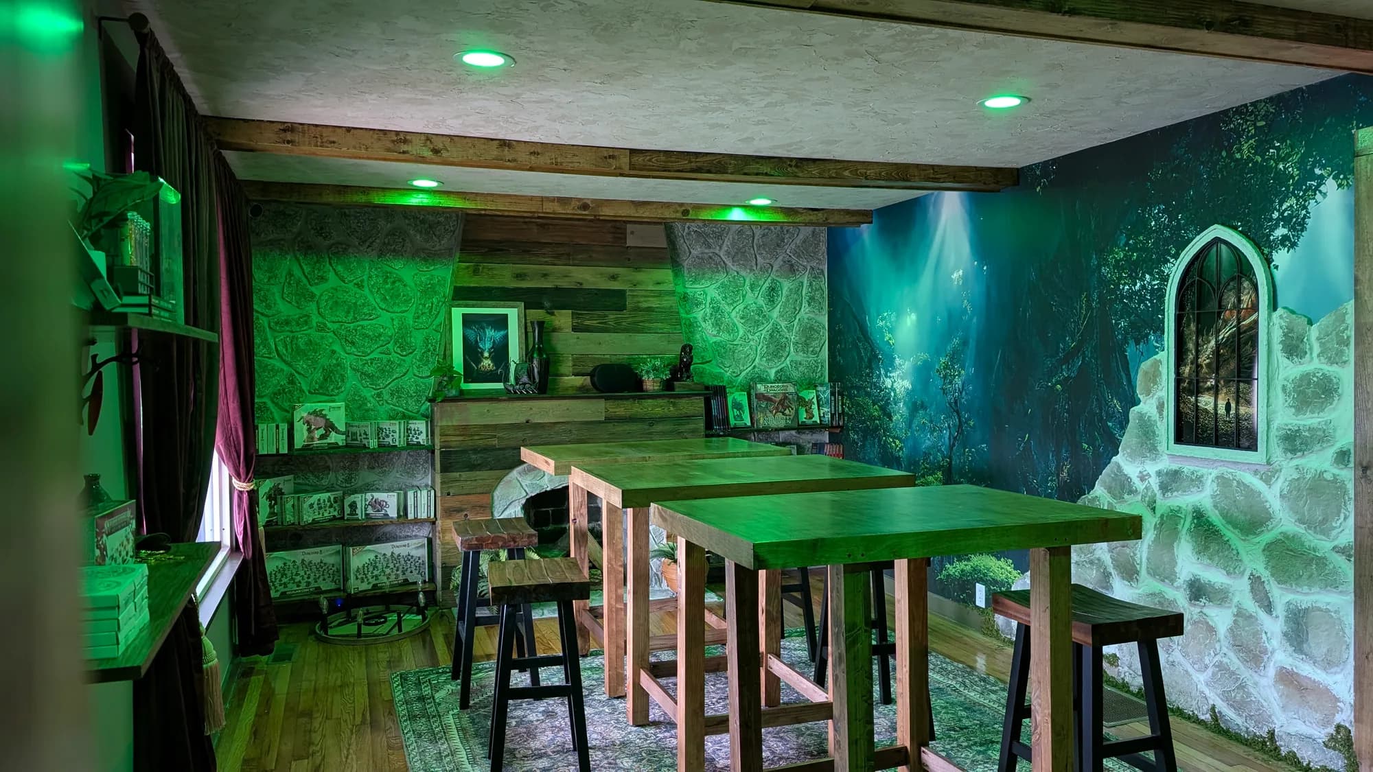 High wooden tables and stools under green light, against a painted forest wall, with rulebooks and boxed miniatures on the shelves behind